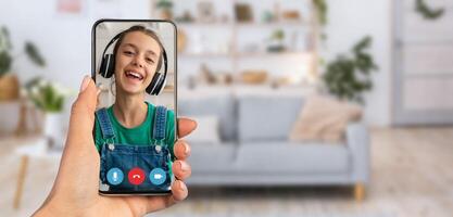 A young child is happily engaging in a call from a smartphone. The setting is a cozy living room with plants and a sofa in the background, creating a warm atmosphere. photo