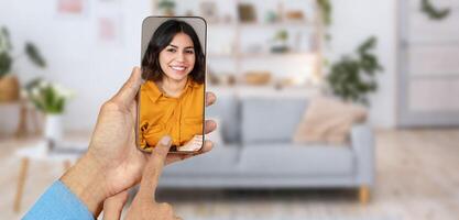 A person engages in an online call using a smartphone, smiling at the screen. The cozy home environment features a sofa and plants, creating a friendly atmosphere for communication. photo