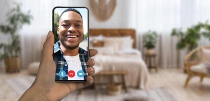 A person is on a call, holding a smartphone with a clear view of a friend's face. The warm and inviting room features soft lighting and plants, creating a comfortable atmosphere. photo