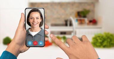 A person is holding a smartphone in their hand during a call. Bright kitchen decor and fresh ingredients in the background create a warm atmosphere for communication. photo