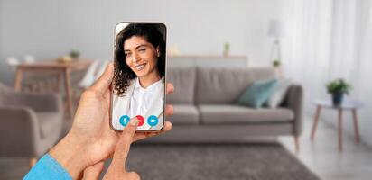 A person engages in a call from home, displaying a screen with smiling face. The setting includes a cozy living room with a sofa and soft furnishings, creating a friendly atmosphere. photo