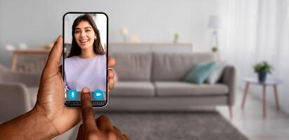 A person holds a smartphone displaying a smiling friend during an online call in a cozy living room. The scene captures modern communication and connection from home. photo