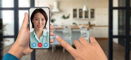 Close-up of a hand holding a smartphone during a call. A woman appears on screen, smiling, while a cozy kitchen setting is visible in the background. The atmosphere feels warm and welcoming. photo