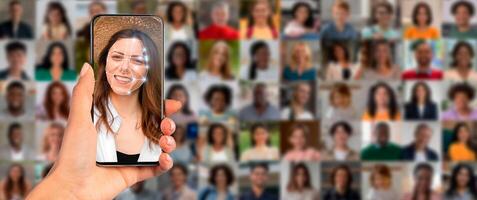 A hand holds a smartphone showcasing facial recognition technology, scanning a smiling woman. In the background, a collage of diverse faces illustrates modern identification methods in action. photo