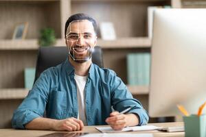A man smiling and sitting at his desk is undergoing facial recognition technology as he participates in a remote meeting. The setting includes bookshelves and a computer monitor. photo