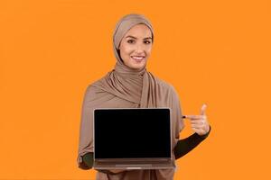 Great Website. Muslim Lady Pointing At Laptop Computer Empty Screen Recommending Internet Service Or Blog Standing Posing On Yellow Studio Background. Mockup photo