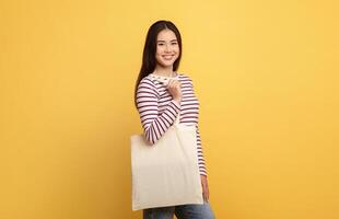 A young Asian woman stands against a cheerful yellow backdrop, smiling while holding a simple tote bag. She wears a stylish striped long-sleeve shirt and casual jeans, radiating positivity. photo