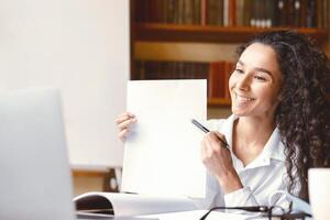 Smiling female manager having videocall on laptop, giving virtual presentation or online lesson to students. Woman showing document contract to webcam, pointing to blank empty sheet of paper photo
