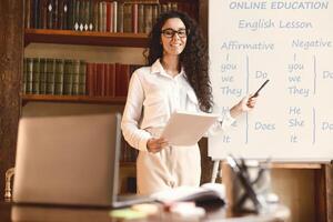 Online Education And E-Learning Concept. Smiling female English teacher standing and pointing at whiteboard with pen, explaining grammar rules. Excited lady wearing glasses and looking at laptop photo