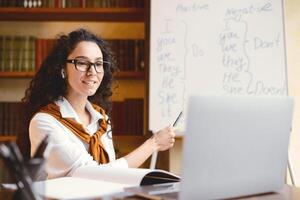 E-learning, Online English Course Concept. Professional female teacher having call, explaining new theme to students, sitting at table and pointing with pen at whiteboard with grammar rules photo