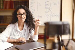Online Teaching And Studying. Professional young woman wearing eyeglasses giving virtual class, sitting at desk, explaining and talking to camera on tripod. Lady filming her internet blog photo