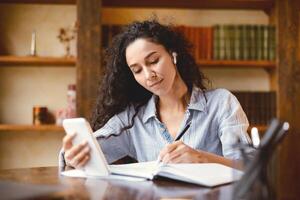 Planning The Day. Young lady in wireless earphones sitting at table, holding and using mobile phone, writing with pen in paper notebook, taking notes about her schedule, watching online course photo