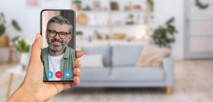 A person holds a smartphone displaying a smiling man on a call. The background reveals a cozy living room environment with a sofa and decor, indicating a relaxed setting for communication. photo