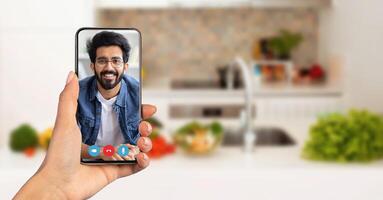 A person holds a smartphone displaying a smiling face during an online call. The background features a well-lit kitchen with fresh vegetables, capturing a moment of connection from home. photo