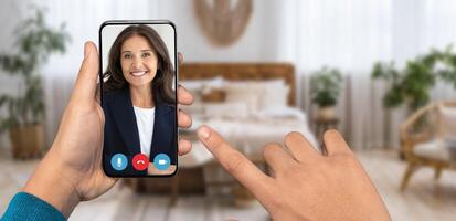 A person is holding a smartphone displaying a smiling face during an online conversation at home. The cozy room features a bed and potted plants, emphasizing a relaxed atmosphere. photo