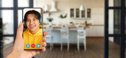 A person is holding a smartphone displaying a smiling individual on a call. The setting is a cozy home with a modern kitchen visible in the background. photo