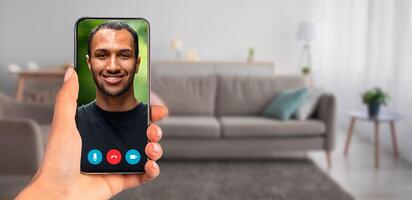 A person holding a smartphone that displays a call with handsome guy. The background features a cozy living room with a couch and natural light, creating a relaxed atmosphere for communication. photo