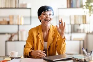 A woman wearing a yellow blazer engages in a virtual conversation, smiling while holding a pen. Facial recognition technology is capturing her features in an office setting. photo