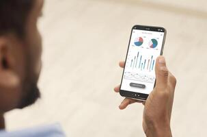Cropped of black man using analytics mobile app on smartphone, checking his incomes on trading stocks, panorama. Male hands holding phone with newest analytics mobile application photo