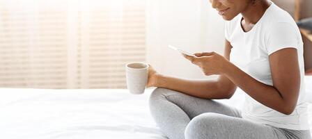 Smiling african american young lady in pajamas drinking morning tea or coffee, using mobile phone, checking social media or reading messages, sitting on bed at home, side view, copy space photo