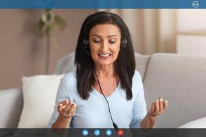 Online Call And Distance Communication. Portrait of senior lady in headphones sitting on sofa at desk, using computer, having virtual conference on laptop talking to webcam, explaining plan photo