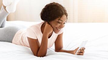 Relaxed african american young lady laying on bed with smartphone, chatting with friends via social media or reading blog, using nice applications, resting on weekend at home, copy space photo