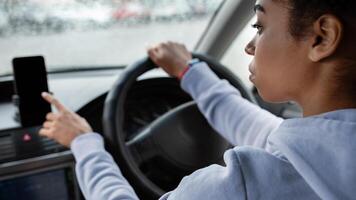 Make route without traffic jams on way to work. Serious young african american woman drives car and use smartphone with empty screen and online map in rainy weather, cropped, side view, copy space photo