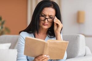 Portrait of mature woman touching glasses trying to read book, having difficulties seeing text because of vision problems. Negative emotion facial expression feelings reaction, health issues photo