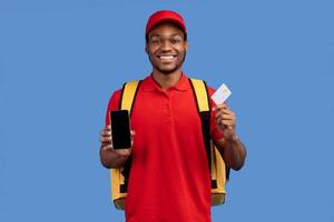 Application For Online Order. Smiling black deliveryman showing credit card and mobile phone with blank screen for mockup, wearing red cap, uniform and thermo backpack bag, isolated on blue background photo