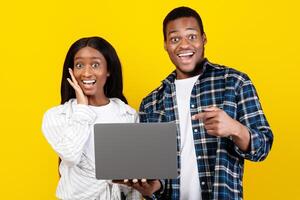 Amazing offer, good site and celebrating victory. Surprised cheerful millennial african american husband and wife hold laptop, man points finger at gadget, isolated on yellow background, studio shot photo