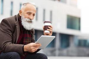 Grey-haired elderly man in warm outfit using digital tablet and drinking coffee to go while sitting on the street, enjoying his free time outdoors, reading book or checking social media, copy space photo