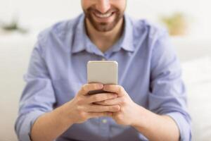 Happy Bearded Guy Using Smartphone Texting And Using Mobile Application On Phone Sitting On Couch At Home. Great App, Man Using Cellphone Browsing Internet Or Playing Game Indoor. Shallow Depth photo