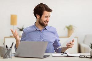 Displeased Businessman Looking At Smartphone Reading Negative Message Sitting At Laptop In Modern Office. Bad News, Mobile Communication Issue, Problems At Workplace Concept. Selective Focus photo