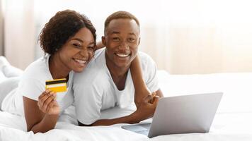 Young black couple laying on bed with laptop, shopping online, recommending credit card, closeup, copy space. Smiling african american man and woman in pajamas making online order, using computer photo