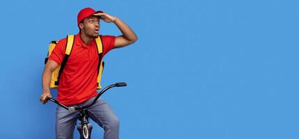 Fast Delivery Service. Emotional surprised black courier guy in red uniform riding bicycle, wearing thermo backpack bag and looking at copy space isolated on blue studio background, banner, panorama photo