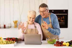 Joyful Senior Couple Calling Using Laptop Waving Hands To Computer, Communicating With Distant Friends Online Standing In Kitchen At Home. Modern Remote Communication, Call Concept photo