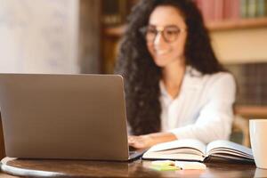 Workspace. Selective focus on notebook on the table, smiling woman in eyeglasses sitting at desk, using laptop in the blurred background. E-learning, remote work, blogger or journalist writing article photo