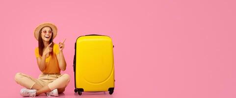 Joyful young lady in summer hat and sunglasses sitting on floor by luggage and pointing at copy space, pink studio background. Happy woman in summer clothes showing exciting travelling deals photo