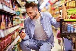 Consumption And Consumerism. Portrait Of Smiling Bearded Man With Shopping Cart In Market Buying Groceries Food Taking Products From Shelves In Store, Holding Glass Jar Of Sauce, Checking Label photo