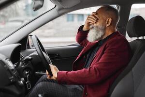 Exhausetd aged grey-haired man having headache while driving car, touching his forehead, side view, shot inside auto, copy space. Elderly businessman having financial crisis or got stuck in traffic photo