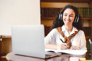 Distance Education. Smiling woman in wireless headset sitting at desk, using laptop and writing in notebook. Lady watching tutorial, lecture or webinar, studying online at home. E-learning photo