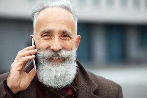 Closeup portrait of cheerful aged man in warm outfit talking on mobile phone while walking by street, copy space. Handsome bearded elderly businessman having conversation on smartphone outdoors photo
