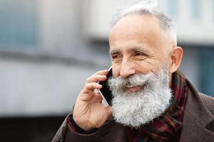 Closeup portrait of cheerful senior man in warm outfit talking on mobile phone while walking by street, copy space. Handsome bearded elderly businessman having conversation on smartphone outdoors photo