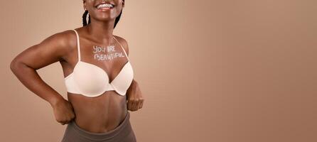 Love Yourself. Cheerful Black Slim Girl With Inscription You Are Beautiful On Chest Posing In Bra Over Beige Background In Studio, Free Space photo