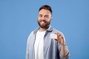 Happy young man showing empty business card with space for design over blue studio background. Smiling bearded guy holding blank advertising card with mockup for your ad photo