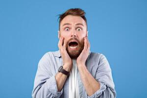 Scared young guy screaming in panic over blue studio background. Horrified bearded man feeling afraid, suffering from panic attack, yelling in fear, being dramatic photo