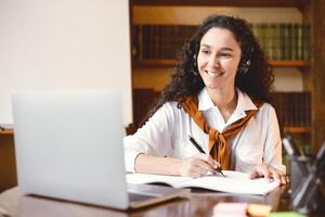 Doing Homework. Portrait of smiling young curly woman sitting at desk with pen, writing and taking notes in notebook, using and looking at laptop. Adult lady watching online course during quarantine photo