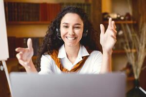 Online Call And Support Concept. Smiling female customer service representative making web conference on laptop computer, talking to camera and consulting client, sitting at table, explaining photo