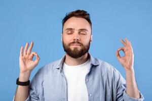 Calm young man meditating to relieve stress, finding inner peace and harmony on blue studio background. Bearded millennial guy practicing mindfulness and deep concentration photo