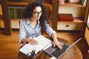 Above top high angle view of casual young woman in eyeglasses working on laptop, typing on keyboard, sitting with open notebook at table in home office or library. Female manager preparing project photo